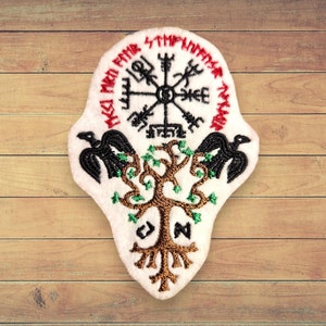 Może przedstawiać: Biała naszywka haftowana z czarnym i złotym drzewem, dwoma czarnymi krukami i czerwono-czarnym kompasem z runami wokół krawędzi. Runy brzmią "HWAET MWAE FAMA", "WYRD" i "WYRD MWAE FAMA".