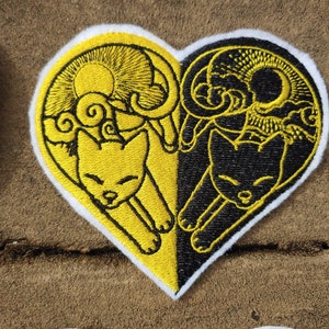 May include: A heart-shaped embroidered patch with two cats, one yellow and one black, with a sun and moon design above them.