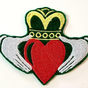 May include: Green felt embroidered patch with a red heart, two white hands holding the heart, and a gold crown on top.