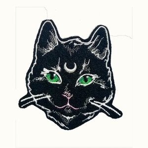 Op de afbeelding: Zwarte geborduurde patch van een kattengezicht met groene ogen en een halve maan boven zijn voorhoofd.
