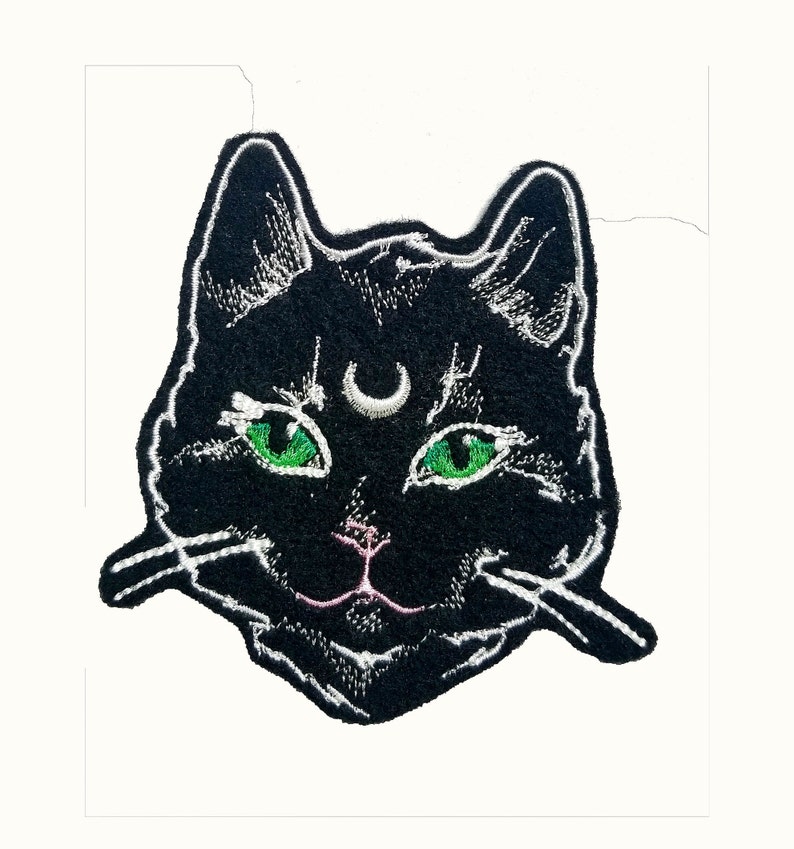 Op de afbeelding: Zwarte geborduurde patch van een kattengezicht met groene ogen en een halve maan boven zijn voorhoofd.