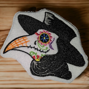 May include: Embroidered patch of a white skull with a black hat and a long orange beak. The skull has colorful sugar skull designs and a black and white patterned beak.