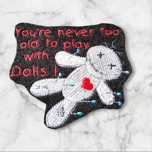 May include: Embroidered patch with a black background and a white doll with a red heart and blue pins. The text "you're never too old to play with dolls!" is embroidered in red.