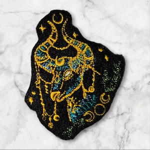 May include: Embroidered iron-on patch with a black background and gold and teal embroidery of a bull's head with a crescent moon and stars.