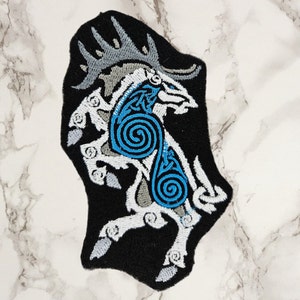 Może przedstawiać: Naszywka do prasowania z haftem przedstawiającym białą, stylizowaną kozę z niebieskim wzorem celtyckim na czarnym tle.