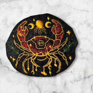 Op de afbeelding: Geborduurde strijkpatch met een zwarte, rode en gouden krab met een zon- en maandesign. De krab is omringd door sterren en bladeren.
