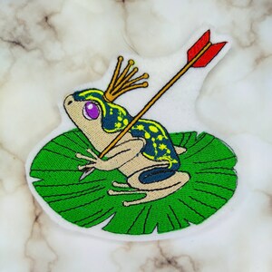 Może przedstawiać: Naszywka haftowana przedstawiająca zieloną żabę w złotej koronie, siedzącą na zielonym liściu lilii wodnej, przez który przechodzi strzała.