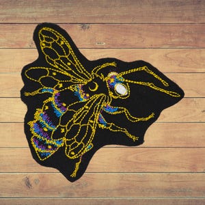 May include: Black embroidered patch of a bee with yellow, blue, and purple details. The bee has a crescent moon and stars on its head.