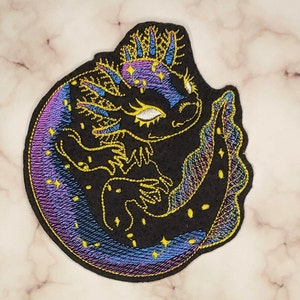 May include: Embroidered patch of a black axolotl with a crown, curled up on a crescent moon with a purple and blue gradient and gold stars.