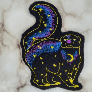 Könnte beinhalten: Schwarzer gestickter Aufnäher mit einem Cartoon-Tier mit einem lila und blauen Galaxieschwanz. Das Tier hat einen Halbmond auf der Brust und gelbe Sterne auf dem Körper.
