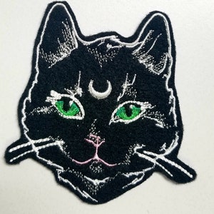 Op de afbeelding: Zwarte geborduurde kattenkop patch met groene ogen en een halve maan op zijn voorhoofd. De patch heeft witte stiksels rond de randen en een roze neus.