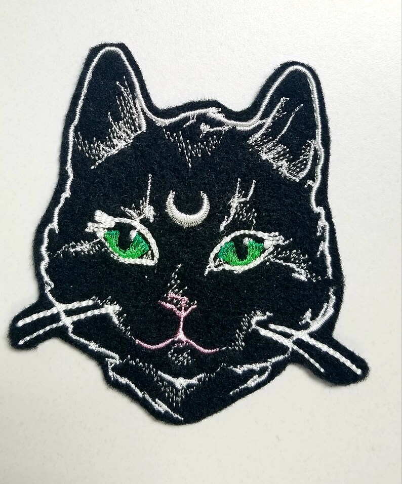 Op de afbeelding: Zwarte geborduurde kattenkop patch met groene ogen en een halve maan op zijn voorhoofd. De patch heeft witte stiksels rond de randen en een roze neus.