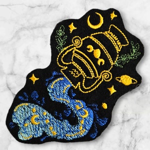 May include: Embroidered iron-on patch with a blue and yellow design of a water pitcher pouring water. The design is on a black felt background and includes stars, moons, and other celestial elements.
