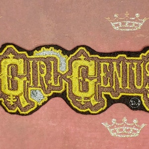 Op de afbeelding: Een geborduurd patch in bruin en goud met het woord "GRRGENIUS" in een gestileerd lettertype.