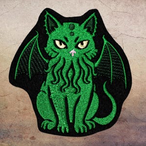 May include: Embroidered patch featuring a green cat-like creature with bat wings and tentacles, set against a black background. The cat has yellow eyes and a pink nose. The patch is designed with detailed stitching.