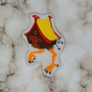 May include: A brown and yellow embroidered patch of a house with legs. The house has a red roof and a yellow roof. The legs are orange and have black claws.