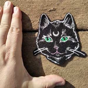 Op de afbeelding: Zwarte geborduurde strijkpatch van een kattengezicht met groene ogen en een halve maan boven zijn hoofd.