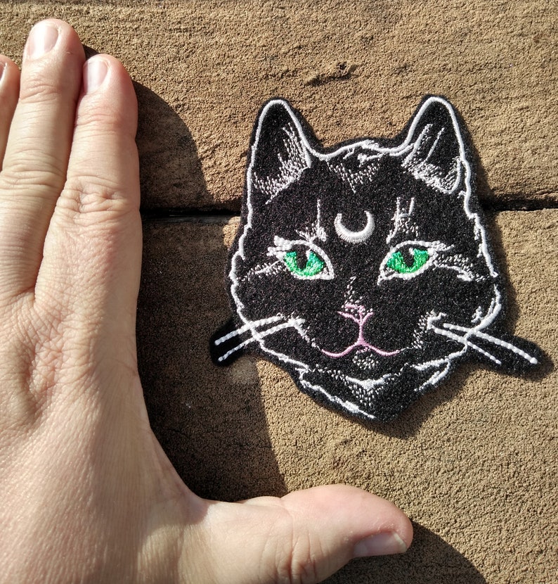 Op de afbeelding: Zwarte geborduurde strijkpatch van een kattengezicht met groene ogen en een halve maan boven zijn hoofd.
