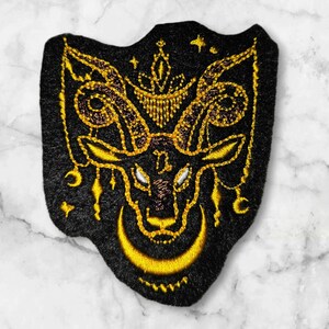 Op de afbeelding: Zwarte geborduurde patch met een gouden rammenkopontwerp. De ram heeft grote hoorns en een halve maan onder zijn kop. De patch is versierd met sterren en andere hemellichamen.