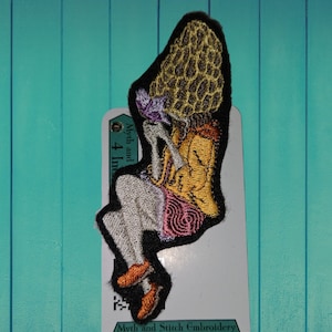 Op de afbeelding: Gestikt patch met een persoon die een grote paddenstoel vasthoudt. De paddenstoel is geel en bruin met een textuur. De persoon draagt een gele en oranje outfit met een roze en paarse rok. Het patch is 10 cm in diameter. De tekst "Myth and Stitch Embroidery" is gedrukt op de witte kaart achter het patch.
