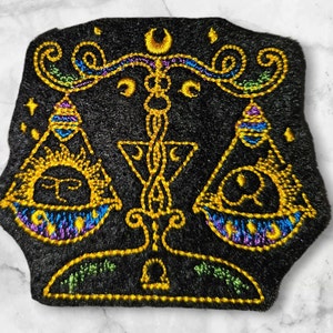 Op de afbeelding: Gestikt patch met een zwarte fluwelen achtergrond met een gouden omtrek van een Libra-sterrenbeeld symbool. De weegschalen zijn versierd met een zon- en maandesign, en het symbool is omgeven door een halve maan en sterren.