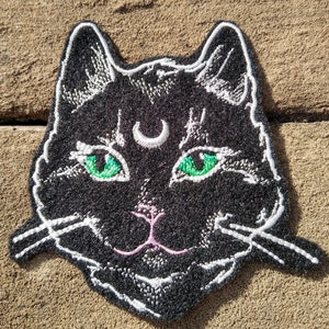 Op de afbeelding: Zwarte geborduurde kattenpatch met groene ogen en een halve maan op zijn voorhoofd. De kat heeft een roze neus en witte snorharen.