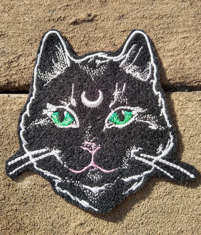 Op de afbeelding: Zwarte geborduurde kattenpatch met groene ogen en een halve maan op zijn voorhoofd. De kat heeft een roze neus en witte snorharen.