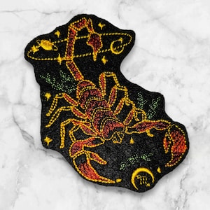 May include: Embroidered iron-on patch featuring a red and gold scorpion with a black background. The scorpion is surrounded by stars and a crescent moon.