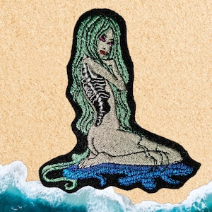 May include: An embroidered patch of a woman with green hair and a skeletal ribcage, sitting on a blue wave. The patch is on a brown corkboard background.