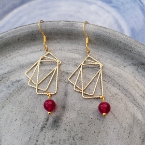 Puede incluir: Pendientes geométricos dorados con una piedra preciosa roja colgante. Los pendientes presentan varios cuadrados superpuestos.
