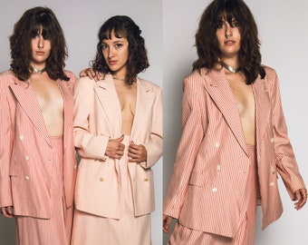 Op de afbeelding: Twee vrouwen dragen bijpassende roze en wit gestreepte dubbelrijdige blazers en rokken. De blazers hebben gouden knopen. De vrouwen staan ​​naast elkaar met hun armen om elkaar heen.