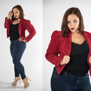 Puede incluir: Una mujer con una chaqueta de gamuza roja, un top negro y pantalones vaqueros azules. Lleva tacones altos y tiene el pelo recogido.