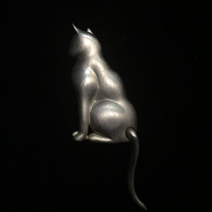 May include: A silver-toned cat brooch with a detailed, realistic design. The cat is sitting with its tail curled around its body.