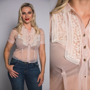 Könnte beinhalten: Eine transparente, hellrosa Bluse mit kurzen Ärmeln und Knopfleiste. Die Bluse hat eine cremefarbene Spitzenblende auf der Brust und am Kragen. Die Knöpfe sind goldfarben.