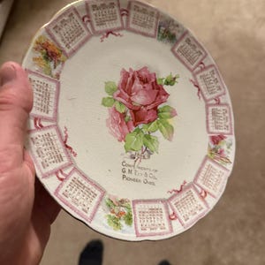 May include: A vintage decorative plate with a pink rose design at the center. The plate features a calendar around the rim and the text "COMPLIMENTS OF G.M. ELY & CO. PIONEER OHIO" and the year 1908.