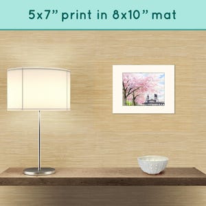 May include: A white lamp with a white lampshade sits on a wooden shelf. A white bowl sits on the shelf to the right of the lamp. A framed print of a watercolor painting of a bridge with pink cherry blossoms hangs on the wall behind the shelf. The print is matted in an 8x10 inch mat. The text "5x7" print in 8x10" mat" is printed above the framed print.