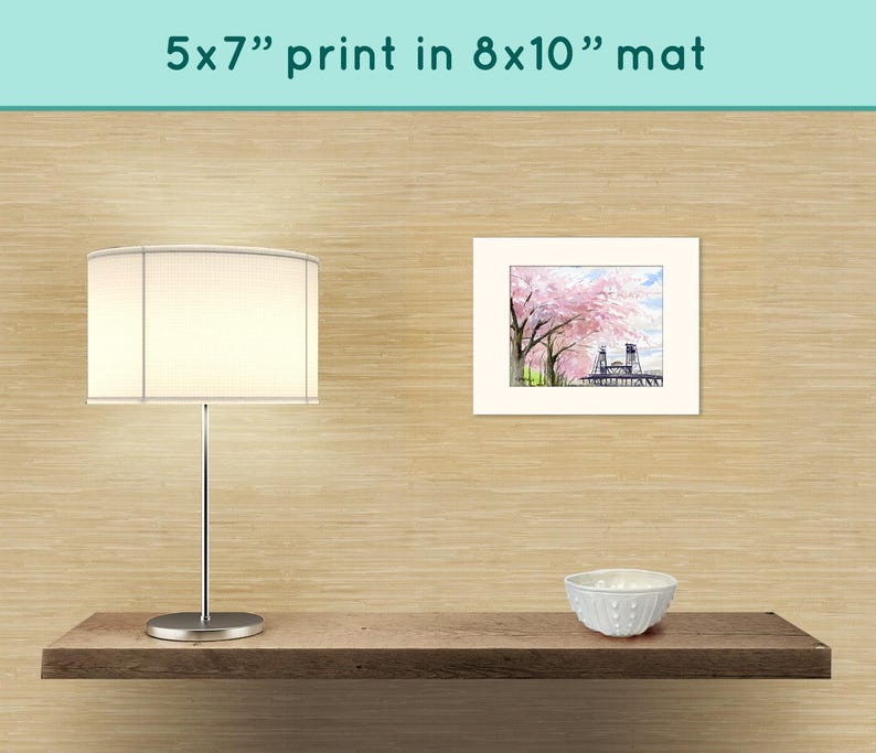 May include: A white lamp with a white lampshade sits on a wooden shelf. A white bowl sits on the shelf to the right of the lamp. A framed print of a watercolor painting of a bridge with pink cherry blossoms hangs on the wall behind the shelf. The print is matted in an 8x10 inch mat. The text "5x7" print in 8x10" mat" is printed above the framed print.