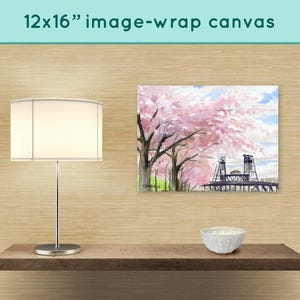 May include: A 12x16 inch canvas print of a watercolor painting depicting a bridge with pink cherry blossoms in the foreground. The canvas is displayed on a shelf with a white lamp and a white bowl.