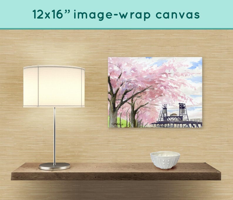 May include: A 12x16 inch canvas print of a watercolor painting depicting a bridge with pink cherry blossoms in the foreground. The canvas is displayed on a shelf with a white lamp and a white bowl.