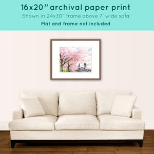 May include: A framed 16x20 inch archival paper print of a watercolor painting depicting a bridge with pink cherry blossoms in bloom. The print is shown above a white sofa.