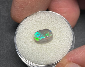 May include: A dark gray oval opal gemstone with green and blue iridescent flashes. The opal is sitting on a white, textured surface.