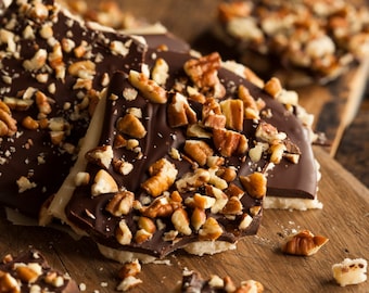 May include: A close-up of a plate of chocolate bark topped with chopped pecans. The bark is broken into irregular pieces and is arranged on a wooden surface.