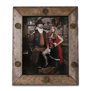 May include: A framed photograph of a couple dressed in Western attire. The man is wearing a brown leather jacket, a white shirt, and a cowboy hat. The woman is wearing a red corset, a black skirt, and a red feather boa. The photo is set in a saloon with a wooden bar and a mirror behind the couple.