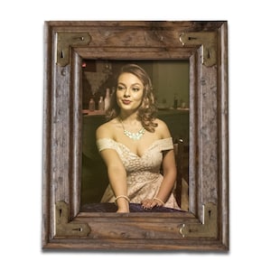May include: A woman in a white dress with a gold necklace sits at a table in a dimly lit room. The image is framed in a dark wood frame with ornate metal corners.