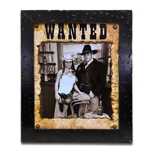 May include: A framed wanted poster with a sepia-toned photo of a couple dressed in western attire. The poster has a distressed, aged look and the text "WANTED" is printed in bold, black letters.
