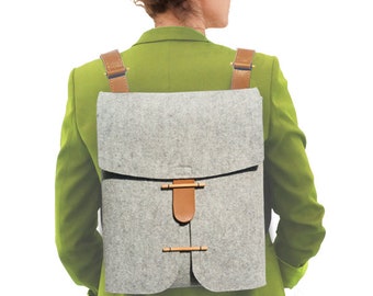 May include: A gray felt backpack with brown leather straps and closures. The backpack has a rectangular shape with a flap closure and adjustable straps. The bag is worn over the shoulders.