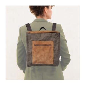 Puede incluir: Una mochila cuadrada con un bolsillo de ante marrón y cierre de cremallera. El cuerpo principal de la bolsa es de color gris oscuro y las correas son negras y grises estampadas. La mochila la lleva una persona con una blazer verde.