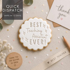 May include: A round, scalloped-edge biscuit with white icing featuring the embossed text BEST Teaching Assistant EVER! surrounded by small star icons. The biscuit is accompanied by a wooden disc with text, a thank you card, and a ribbon.