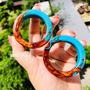 May include: Two resin bangles with a blue and orange marbled design. The bangles are stacked on top of each other and have a gold fleck throughout.
