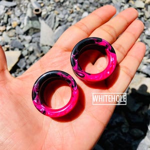 Puede incluir: Dos expansores circulares para orejas con un vibrante diseño veteado en rosa y negro. Los expansores se sostienen en una mano, mostrando los colores brillantes y el patrón único. El texto "WHITEHOLE" es visible.